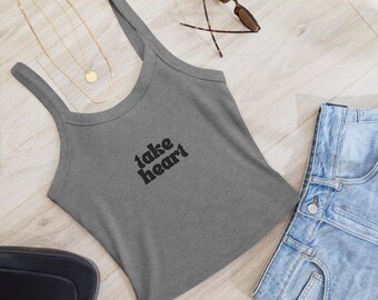 May include: A gray tank top with the text "take heart" printed in black on the front. The tank top is laying on a wooden surface with a pair of sunglasses and a pair of blue jeans.