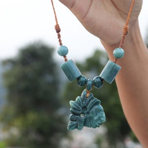 Puede incluir: Un collar turquesa con un colgante tallado con un diseño de rostro. El collar tiene cuentas cilíndricas y esféricas, ensartadas en un cordón marrón. El fondo está borroso, con árboles verdes y un cielo nublado.