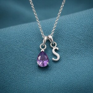 May include: A silver necklace with a delicate chain. The pendant features a pear-shaped, purple gemstone and a silver letter "S". The necklace is displayed on a teal blue fabric.