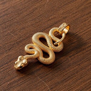 Op de afbeelding: Een goudkleurige slangenhanger met een getextureerd oppervlak. De slang is opgerold en heeft een gedetailleerde kop en staart. De hanger heeft een lus voor een ketting en ligt op een houten oppervlak.