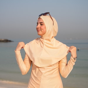 May include: A woman wearing a peach-coloured long-sleeved swimsuit with a matching headscarf. She is standing on a sandy beach with the sea in the background.