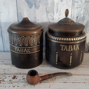 Puede incluir: Dos tarros de tabaco vintage de cerámica marrón con detalles dorados y la palabra "TABAK" en la parte delantera. Una pipa de madera marrón está en primer plano.