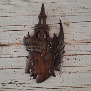 Op de afbeelding: Een donkerbruine houten wanddecoratie met een gegraveerd gezicht en een gedetailleerde hoofdtooi. Het gezicht heeft een serene uitdrukking en de hoofdtooi is ingewikkeld en versierd.