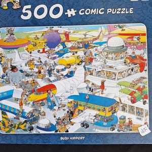 Puede incluir: Un rompecabezas c&oacute;mico de 500 piezas que representa un aeropuerto concurrido. La imagen muestra numerosos aviones, veh&iacute;culos y personas en un estilo colorido y de dibujos animados. El texto "BUSY AIRPORT" es visible en la parte inferior.