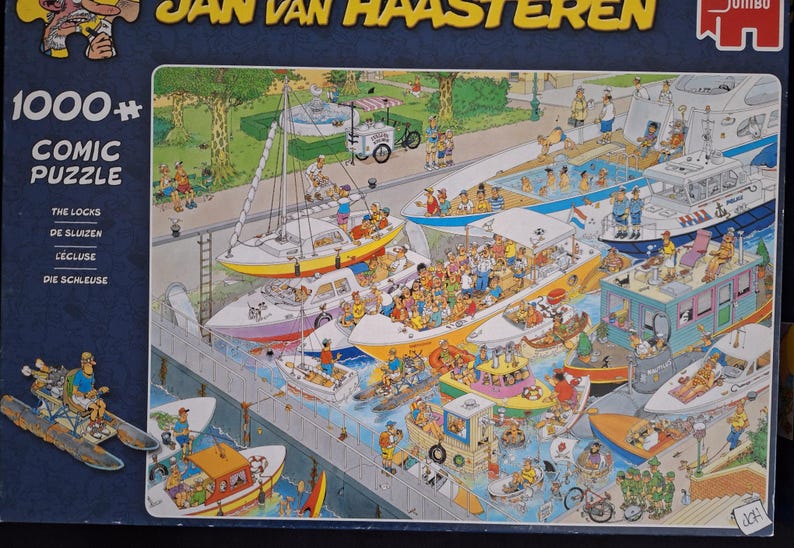 May include: A 1000+ piece comic puzzle by Jan van Haasteren. The image shows a bustling harbour scene with many boats, people, and a food cart. The puzzle box has text in multiple languages, including English, Dutch, and French.