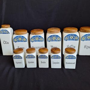 Könnte beinhalten: Set aus 11 weißen Keramikbehältern mit blauen und gelben Blumendesigns und Holzdeckeln. Die Behälter sind mit niederländischen Wörtern für verschiedene Zutaten beschriftet, darunter "Olie" (Öl), "Thee" (Tee), "Koffie" (Kaffee) und "Rijst" (Reis).