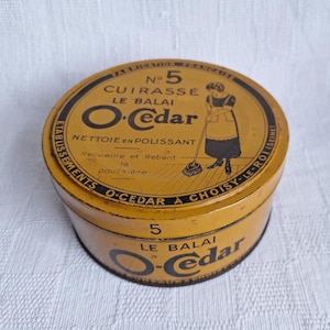 May include: A yellow metal tin with black lettering and a black and white illustration of a woman sweeping. The text on the tin reads "No. 5 CUIRASSÉ LE BALAI O-Cedar NETTOIE EN POLISSANT Recueille et Retient la poussière ETABLISSEMENTS O-CEDAR A CHOISY-LE-ROI (SEINE) 5 LE BALAI O-Cedar".
