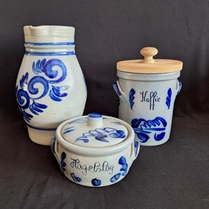 Op de afbeelding: Een keramische kan en twee voorraadpotten met deksel, voorzien van blauwe handgeschilderde bloemmotieven op een lichtgrijze ondergrond. Eén pot is gelabeld Koffie en de andere Hagelslag, met zijhandvatten en een lichthouten deksel op de hoge pot.