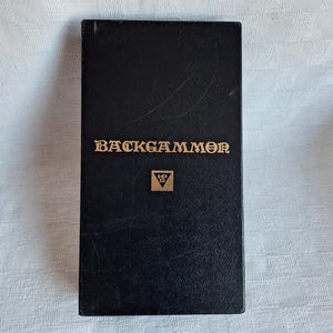 Peut inclure: Boîte noire avec le texte "BACKGAMMON" en lettres dorées et un logo triangle doré.