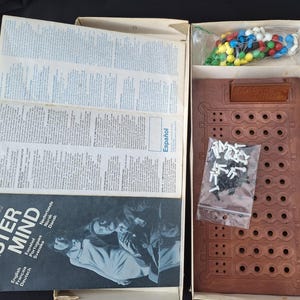 May include: Open box of the board game Master Mind. The game board is brown with multiple holes. The box contains colored pegs, black and white pegs, and instruction manuals. The game's title is printed on the box.