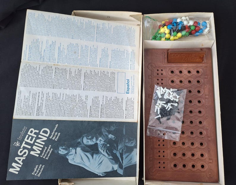 May include: Open box of the board game Master Mind. The game board is brown with multiple holes. The box contains colored pegs, black and white pegs, and instruction manuals. The game's title is printed on the box.