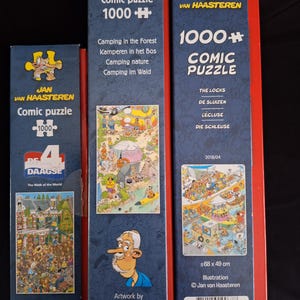 May include: Three Jan van Haasteren comic puzzle boxes. The boxes are red, blue, and white. The boxes have the text "Comic puzzle 1000+" and feature illustrations of camping and other scenes. The boxes are from the Jumbo brand.