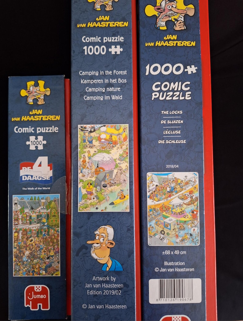 May include: Three Jan van Haasteren comic puzzle boxes. The boxes are red, blue, and white. The boxes have the text "Comic puzzle 1000+" and feature illustrations of camping and other scenes. The boxes are from the Jumbo brand.