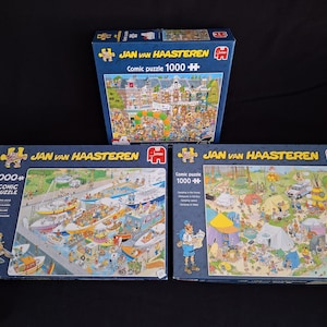 May include: Three Jan van Haasteren 1000-piece comic puzzles. The boxes are blue with detailed illustrations of busy scenes. The puzzles feature drawings of people, boats, and camping activities. The text "Comic puzzle 1000+" is visible on the boxes.