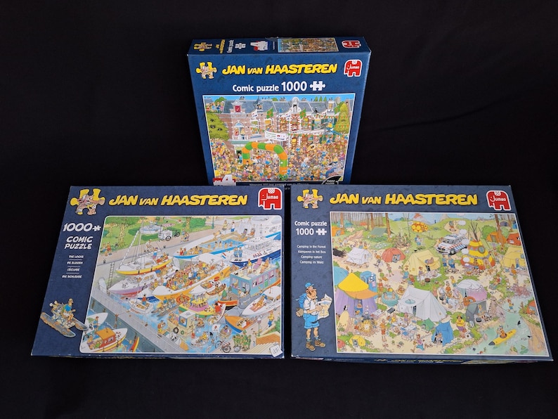 May include: Three Jan van Haasteren 1000-piece comic puzzles. The boxes are blue with detailed illustrations of busy scenes. The puzzles feature drawings of people, boats, and camping activities. The text "Comic puzzle 1000+" is visible on the boxes.