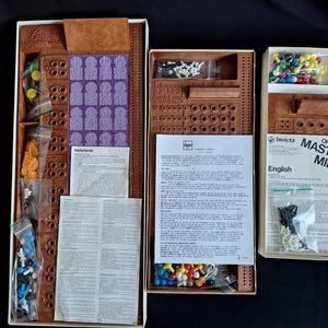 May include: Open boxes of the board game Master Mind, featuring brown game boards, colored pegs, and instruction sheets. The game pieces are in clear plastic bags. The box on the right has the words "Original Master Mind" printed on it.