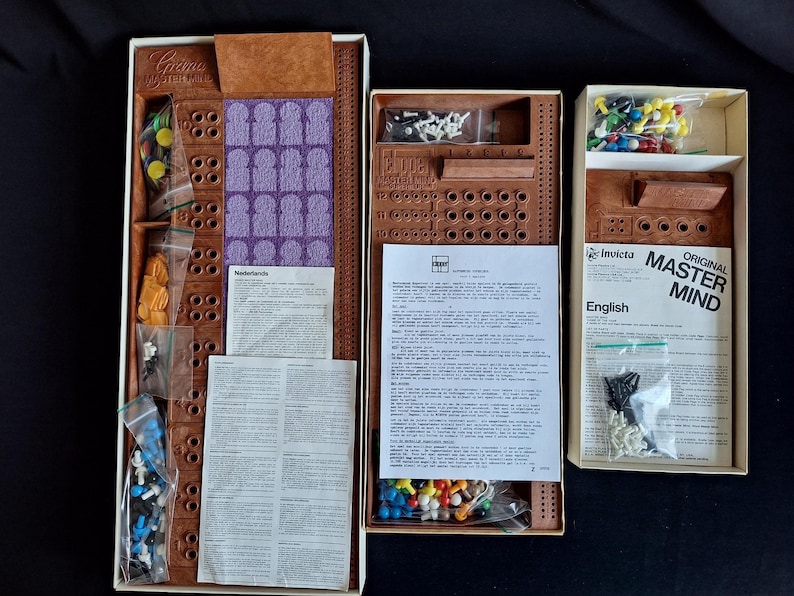 May include: Open boxes of the board game Master Mind, featuring brown game boards, colored pegs, and instruction sheets. The game pieces are in clear plastic bags. The box on the right has the words "Original Master Mind" printed on it.