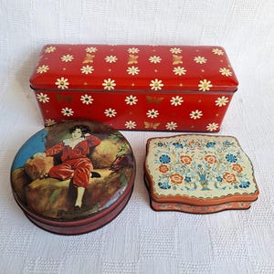 Op de afbeelding: Drie vintage blikken dozen met verschillende ontwerpen. Een doos is rood met witte madeliefjes en gouden vlinders. Een andere doos is rond met een kleurrijk beeld van een vrouw in een rode jurk. De derde doos is vierkant met een bloemmotief in tinten blauw, oranje en groen.