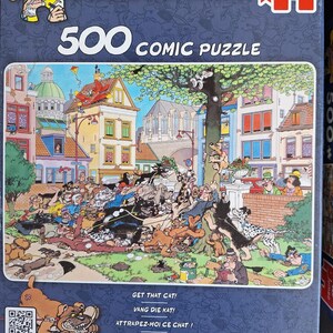 Puede incluir: Una caja de rompecabezas c&oacute;mico de 500 piezas con una colorida ilustraci&oacute;n de una escena bulliciosa con numerosos perros en una plaza de la ciudad europea. La caja incluye el texto "500 COMIC PUZZLE" y las frases "GET THAT CAT!", "VANG DIE KAT!" y "ATTRAPEZ-MOI CE CHAT!"