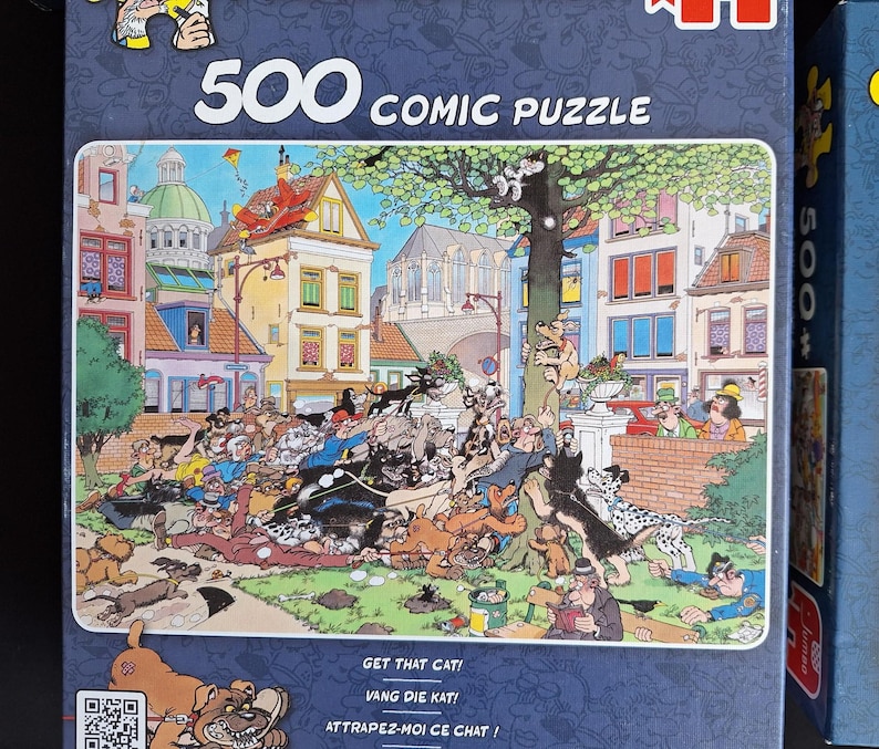 Puede incluir: Una caja de rompecabezas c&oacute;mico de 500 piezas con una colorida ilustraci&oacute;n de una escena bulliciosa con numerosos perros en una plaza de la ciudad europea. La caja incluye el texto "500 COMIC PUZZLE" y las frases "GET THAT CAT!", "VANG DIE KAT!" y "ATTRAPEZ-MOI CE CHAT!"