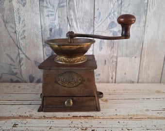 May include: An antique coffee grinder with a wooden base and a brass bowl. The grinder has a hand crank with a wooden handle and a small drawer. The overall color is a mix of brown and gold, with a vintage aesthetic.