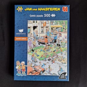 Op de afbeelding: Een rechthoekige Jan van Haasteren 500-stukjes strip-puzzeldoos met een kleurrijke, drukke straatscène. De blauwe doos bevat de tekst Jan van Haasteren, Comic puzzle 500, Kinderpostzegels special edition en het Jumbo-logo.
