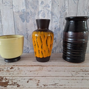 Peut inclure: Trois vases en céramique de styles et de couleurs différents. Le vase de gauche est jaune avec une base noire. Le vase du milieu est brun avec des rayures orange et noires. Le vase de droite est noir avec une bande brune.