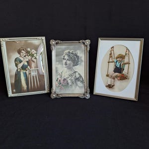 May include: Three rectangular picture frames containing vintage-style portraits. The left frame is silver with a floral print, the centre frame is ornate silver with a portrait of a person, and the right frame is silver with a white oval mat.
