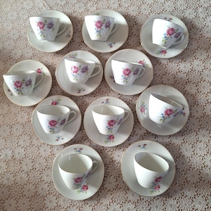 Può includere: Un set di dieci tazze da tè e piattini con motivi floreali. Ogni tazza e piattino presenta un diverso disegno floreale con fiori rosa, viola e bianchi.