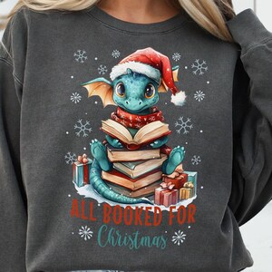 Pode incluir: Uma camisa de moletom cinza escuro com um design de Natal que apresenta um dragão azul usando um chapéu de Papai Noel e lendo um livro. O dragão está sentado em uma pilha de livros e cercado por presentes. O texto "All Booked For Christmas" está impresso abaixo do dragão.