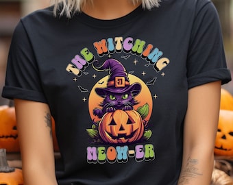 Op de afbeelding: Zwart T-shirt met een afbeelding van een zwarte kat met een heksenhoed die een jack-o'-lantern vasthoudt. De tekst "The Witching Meow-er" is in regenboogkleuren gedrukt.