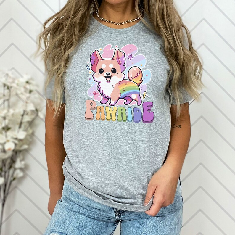 Puede incluir: Una camiseta gris con un perro de dibujos animados que lleva una cola de arco&iacute;ris y el texto "PAWride" en letras de arco&iacute;ris.