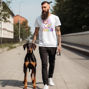 Puede incluir: Un hombre lleva una camiseta blanca con un perro de dibujos animados y el texto "Pawrade" impreso en ella. Camina con un perro negro y marr&oacute;n con una correa.