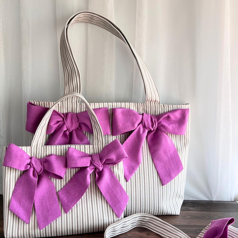 May include: Two tote bags made of beige and white striped fabric, each featuring two large, vibrant purple fabric bows attached to the front. The bags have matching striped shoulder straps and a rectangular shape with a structured design.