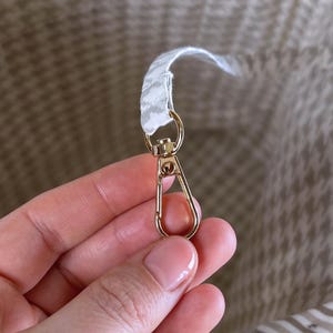 May include: A gold-toned metal lobster clasp attached to a white and grey patterned fabric strap inside a bag. The interior of the bag features a light-colored fabric lining with a subtle houndstooth pattern and horizontal stripes on the side.