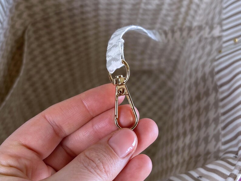 May include: A gold-toned metal lobster clasp attached to a white and grey patterned fabric strap inside a bag. The interior of the bag features a light-colored fabric lining with a subtle houndstooth pattern and horizontal stripes on the side.