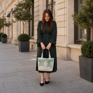 May include: A dark green long-sleeved midi dress with a button-down front and a tie waist, paired with a cream-colored tote bag featuring thin vertical stripes and two large, light green decorative bows attached to the front panel.