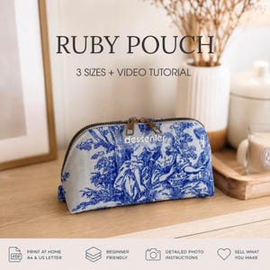 May include: A zippered pouch featuring a blue and white toile de jouy pattern with a landscape scene. The pouch has a rectangular base, a curved top, and a small label with the text "dessenler" attached to the metal zipper pull.