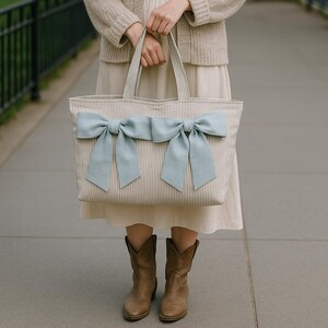 Könnte beinhalten: Eine rechteckige Tragetasche mit einem beigen und weißen vertikalen Nadelstreifenmuster. Die Vorderseite der Tasche ist mit zwei großen, hellblauen Stoffschleifen verziert, die nebeneinander angebracht sind. Die Tasche hat zwei passende Schulterriemen.