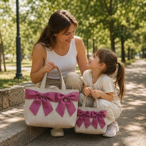 May include: Two beige and white striped tote bags featuring two large, vibrant magenta bows on the front. Each bag has two sturdy top handles and a rectangular structure. The bags are made of a textured fabric and display a consistent design.