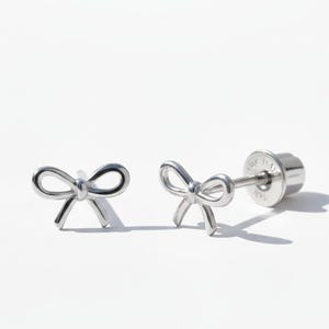 May include: A pair of silver-colored bow-shaped stud earrings. One earring is in focus, showing the post and backing. The other earring is slightly out of focus. The earrings are on a white background.