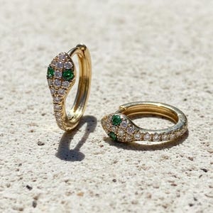 May include: Two gold-colored hoop earrings, each shaped like a snake. The snake heads are adorned with small, clear gemstones and two green gemstones for eyes. The earrings are set against a textured, light-colored surface.