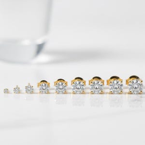 May include: A collection of gold stud earrings with clear, round-cut gemstones. The earrings are arranged in a row, showcasing a range of sizes. The gold settings provide a warm contrast to the sparkling stones, creating a classic and elegant look.