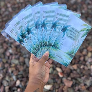 May include: A hand holds a fan of clear plastic sheets with a tropical design. Each sheet has a palm tree illustration and text that reads "52 Week Scripture Savings Challenge" and weekly prompts. The background is a blurred view of small rocks.