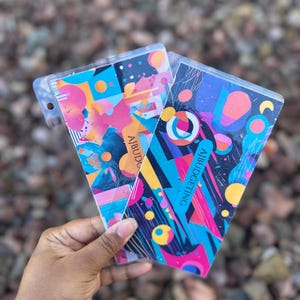 Op de afbeelding: Twee notitieboekjes met abstracte ontwerpen in een transparante plastic hoes. De notitieboekjes hebben een levendige mix van roze, blauwe, oranje en gele geometrische vormen en patronen. De tekst "AJBUDGETING" is zichtbaar.