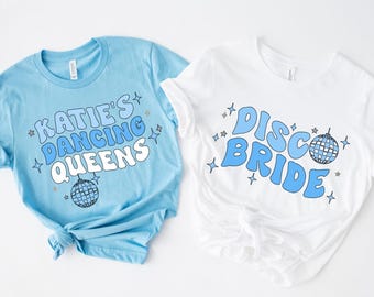 May include: Two t-shirts, one light blue and one white, with retro-style text and disco ball graphics. The light blue shirt says "KATIE'S DANCING QUEENS". The white shirt says "DISCO BRIDE".