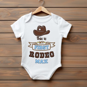 Pode incluir: Macac&atilde;o branco para beb&ecirc; com um chap&eacute;u de cowboy marrom, uma fita azul com o texto "This is my first rodeo Max" em letras marrons e azuis.