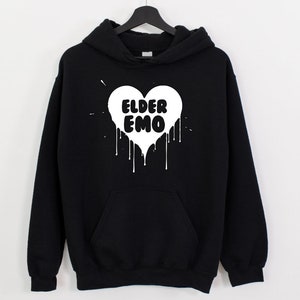K&ouml;nnte beinhalten: Schwarzer Kapuzenpullover mit einem wei&szlig;en Herz-Motiv, auf dem "Elder Emo" in einem tropfenden Farbstil steht.