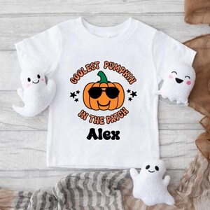 May include: White t-shirt with a pumpkin wearing sunglasses and the text "Coolest Pumpkin in the Patch" with the name "Alex" printed below. Two white ghost plushies are on either side of the shirt.