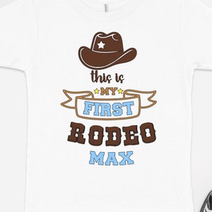 Pode incluir: Camiseta branca com um chap&eacute;u de cowboy marrom, uma faixa marrom com o texto "This is my first rodeo Max" em letras amarelas, azuis e marrons.